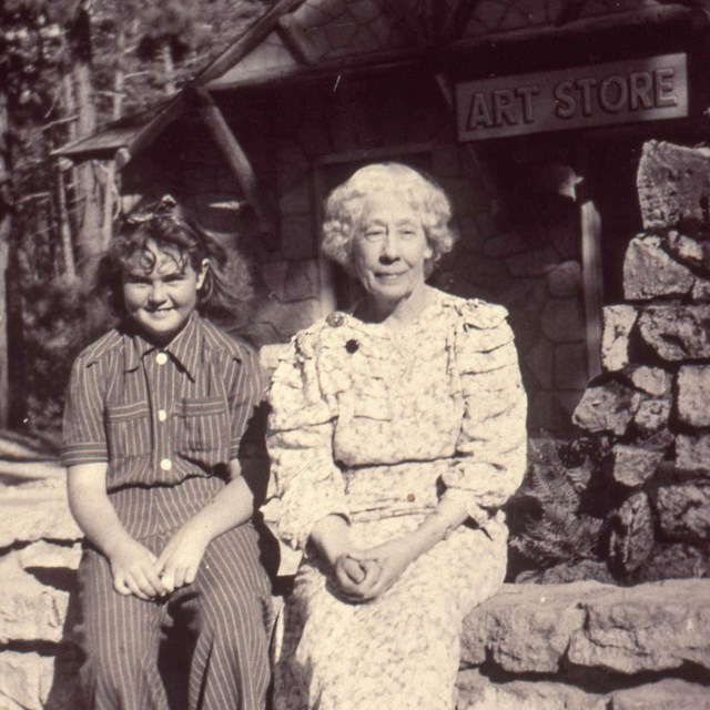 A young girl and older woman sit on a rock wall backed by a building with a sign 