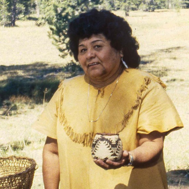 A woman in an animal hide shirt holds a woven basket in front of a meadow.