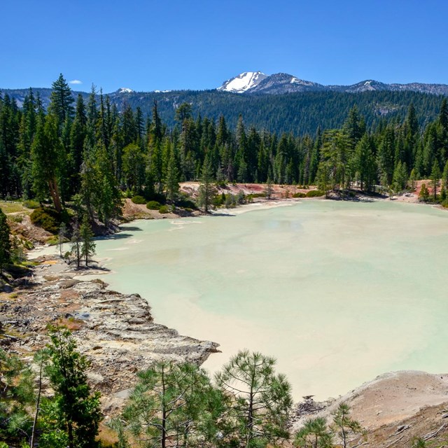 A murky teal lake surrounded by barren soil dotted with conifers backed by a snow-capped peak.