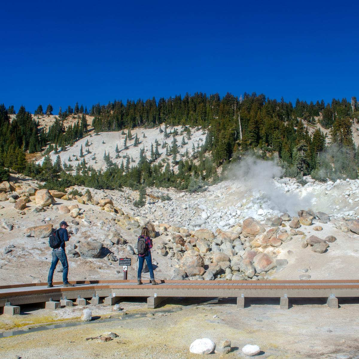 A man and a woman walk along a boardwalk in a hydrothermal area. 