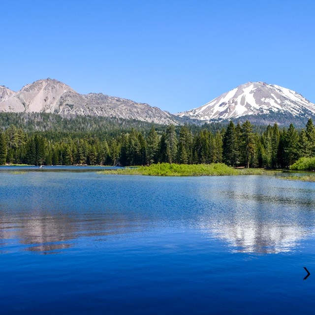 A color photograph of a blue lake lined by conifer forests and large volcanic peaks.