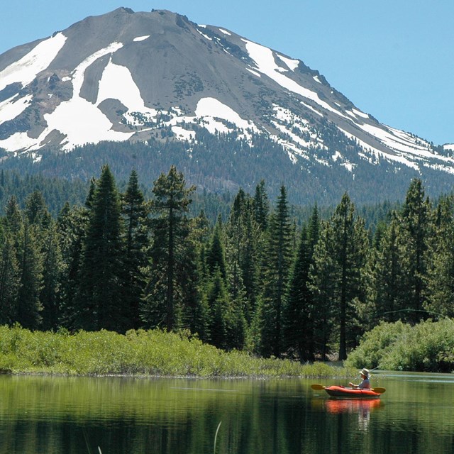 A person holds a fly fishing rod in an orange boat on a lake below a large mountain.