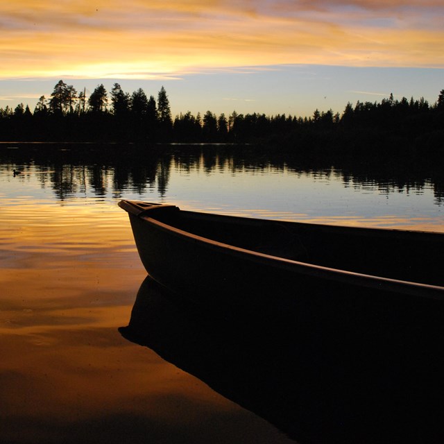 Half of a canoe in a lake lined by conifer trees under an orange sunset sky