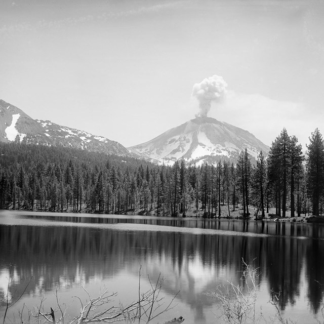 A volcano with a smoke plume fronted by a lake