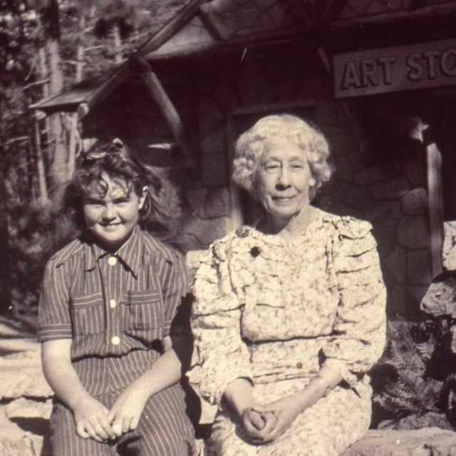 A young girl and older woman sit on a rock wall backed by a building with a sign 