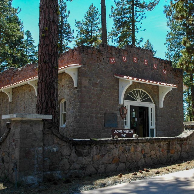 A stone building surrounded by a waist-high stone wall and large conifers.