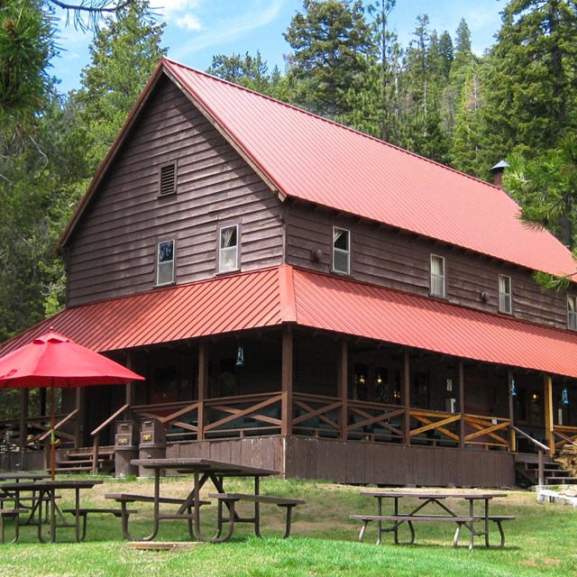 A two-story brown, wooden with a red roof fronted by a grass lawn and surrounded by conifers.