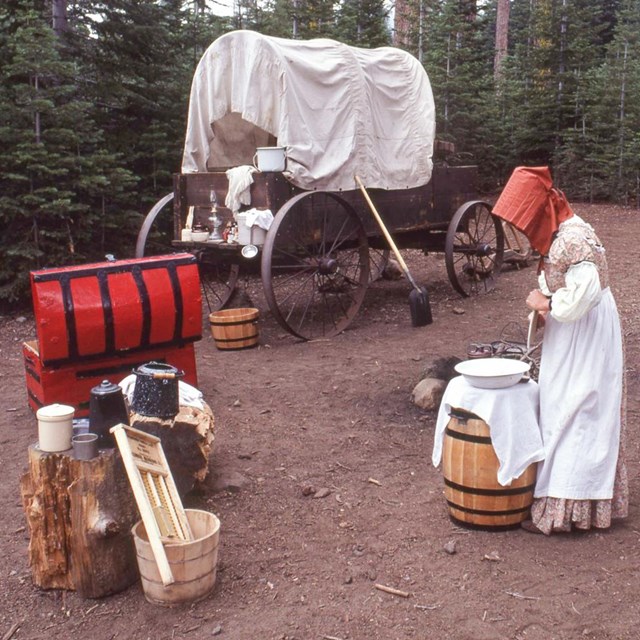 A woman in pioneer costume stands at a barrel near a covered, wooden wagon