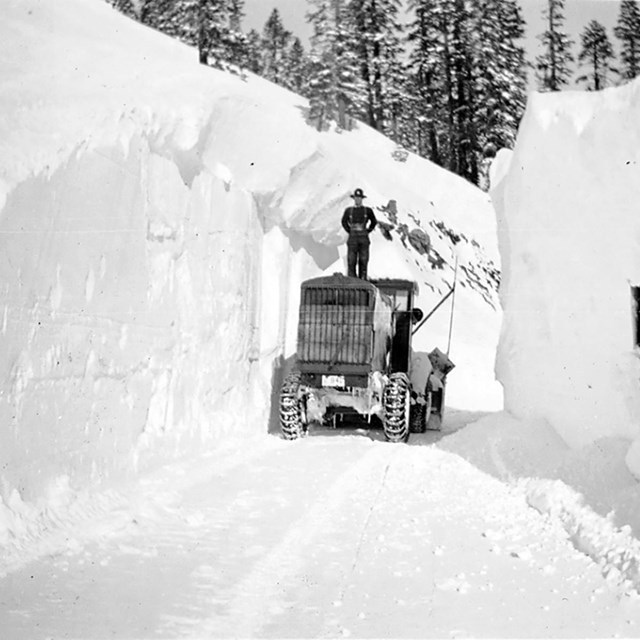 A man stands on a snowplow flanked by walls of snow