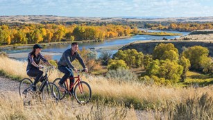 Two Cyclists bike along river with golden fall colors and mountains beyond.