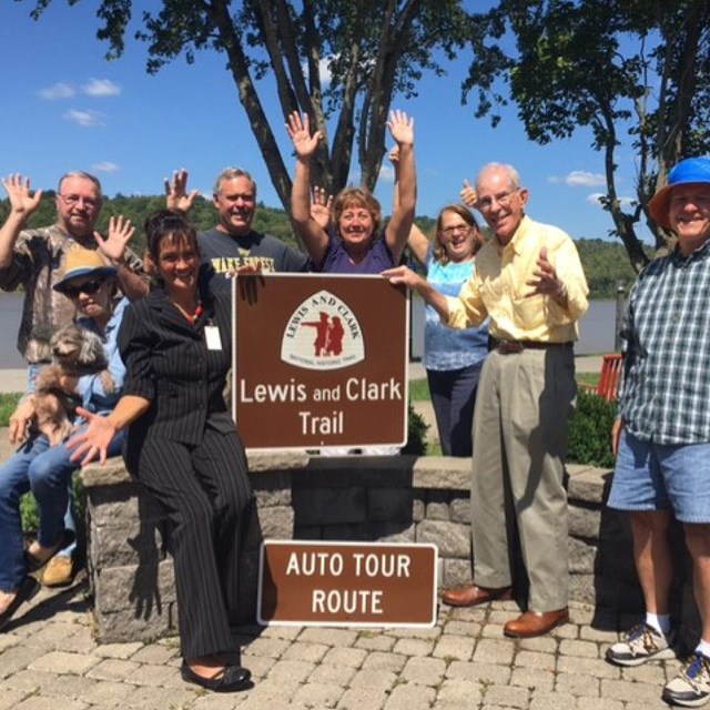 Group poses around brown road sign. Sign reads. Lewis and Clark Trail Auto Tour Route