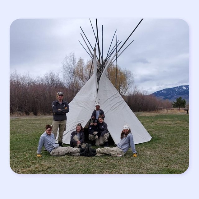 7 young people in AmeriCorps uniforms pose in front of teepee. Mountain behind.