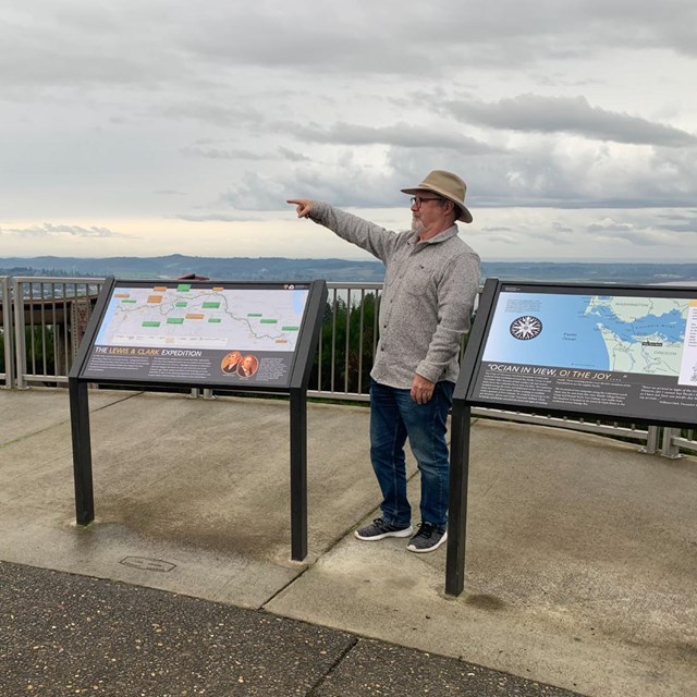 Man poses with two new Lewis and Clark Trail waysides. Evergreen hills behind.