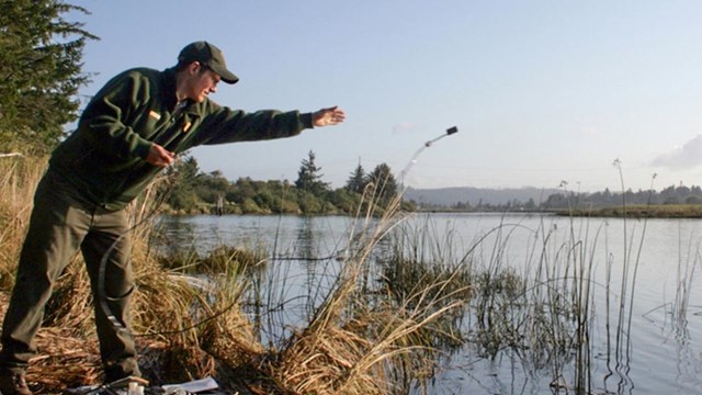 A person in NPS uniform tosses a small device connected to rubber tubing out into a body of water