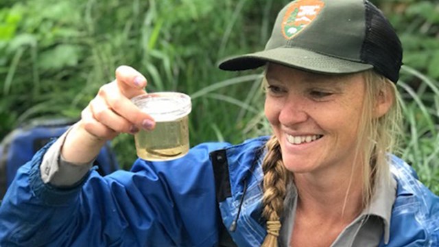 A woman in an NPS ballcap holding a plastic container with an aquatic organism in it