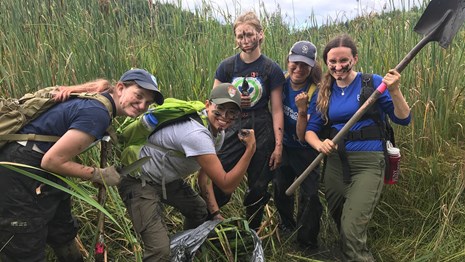 Five women with muddy faces, in wetlands, carrying tools and a bag of pulled plants.