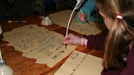 A student writes with a quill pen on parchment in a wooden hut.