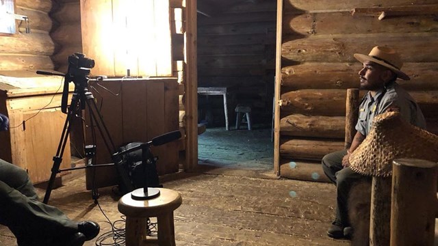 A camera filming a ranger sitting on a bed in the captains quarters of Fort Clatsop