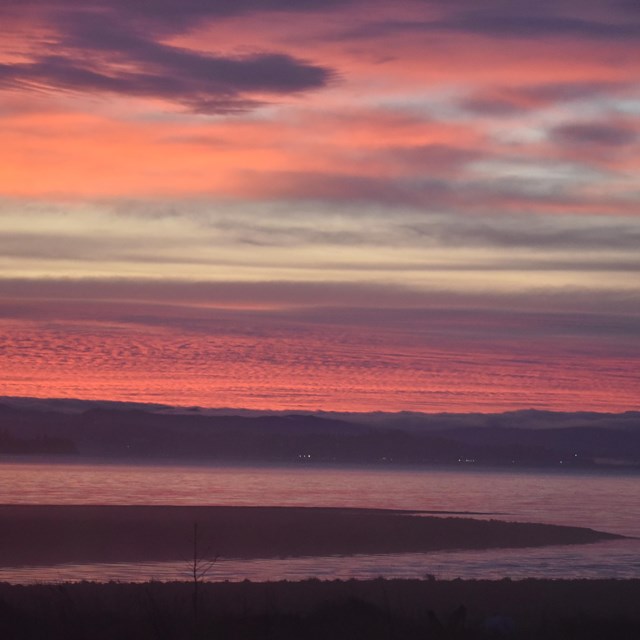 Sunset over a body of water. Clouds and water are streaked pink and purple due to smoke. 