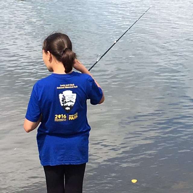 The back of a young girl in blue tshirt as she holds a fishing pole and looks out over the water.