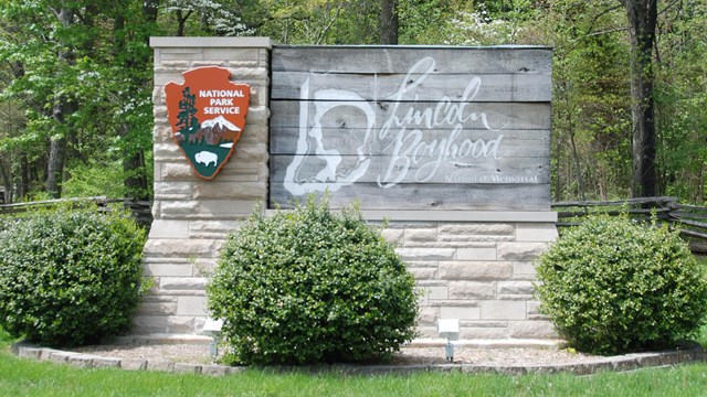 Stone and wood entrance sign surrounded by shrubs, trees and green grass.