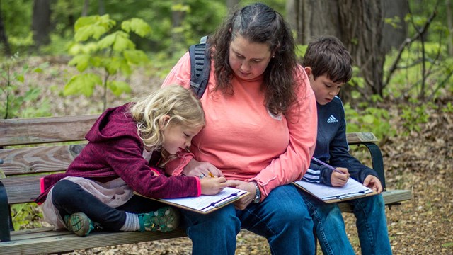 woman and two kids sit on bench on park trail writing in a junior ranger book, trees in background