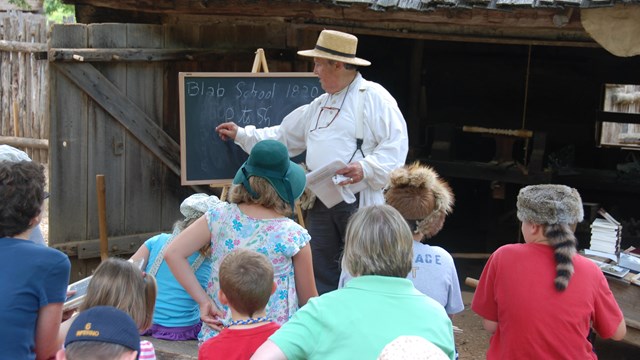 Man in pioneer clothing writing on chalkboard teaching 8 young kids