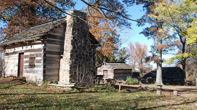 Log cabin and smokehouse surrounded by trees with brown leaves and blue sky