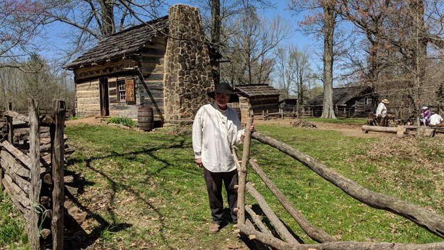 Man in pioneer clothing standing by wooden gate, log cabin and outbuilding in background