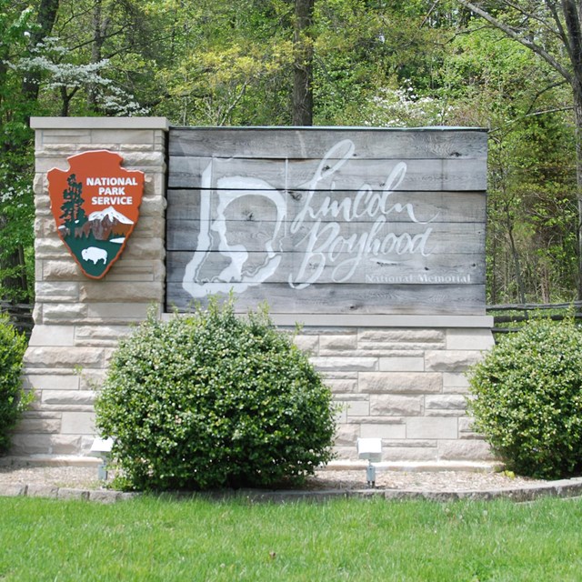 Stone and wood entrance sign surrounded by shrubs, trees and green grass.