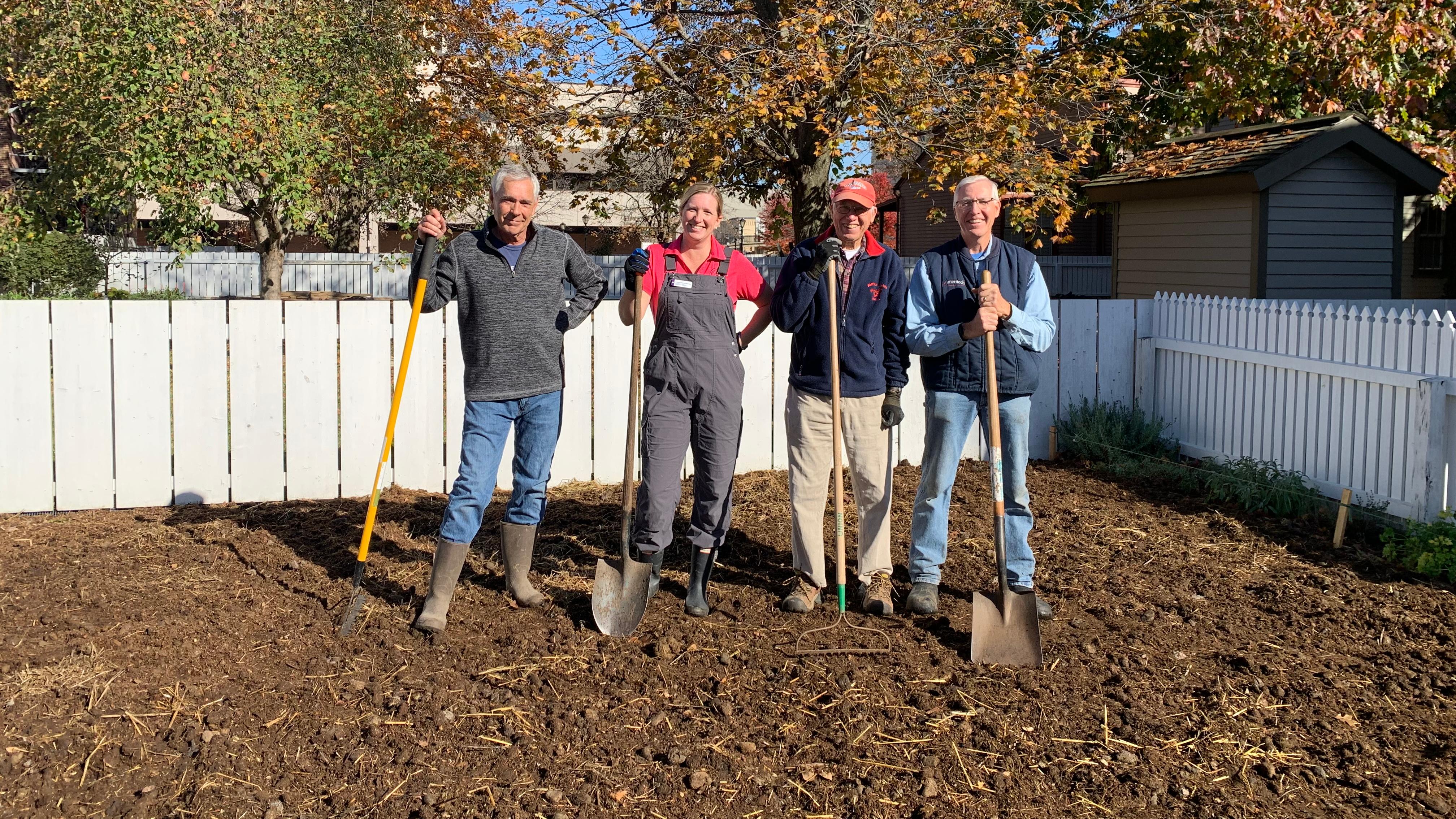  A group of volunteers holding shovels and standing in a fenced-in empty garden.