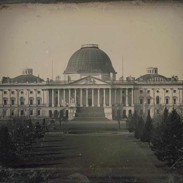 U.S. Capitol in 1846: long, white 3 story building with 3 dark domes, the middle being the tallest.