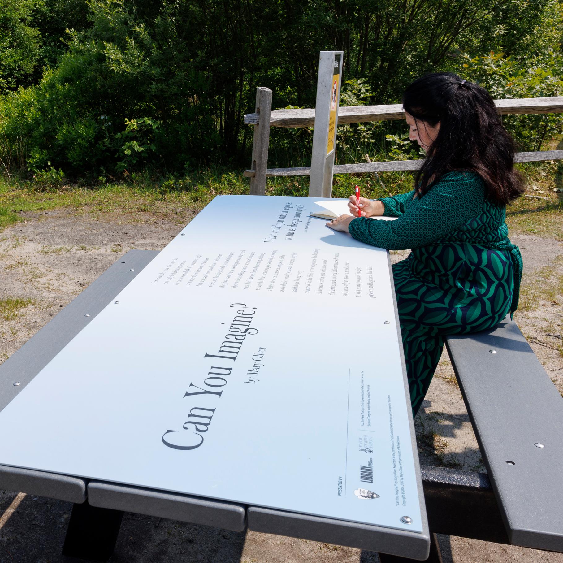 A person writes at a picnic table covered with a panel featuring a poem.