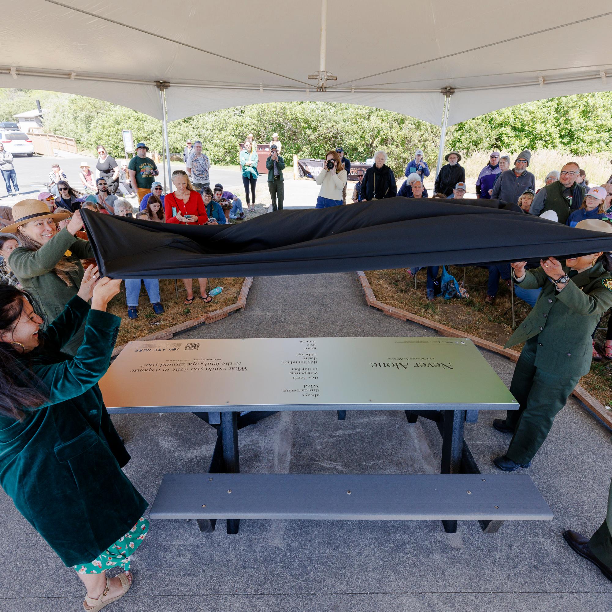 Four people unveil a picnic table featuring a poem.