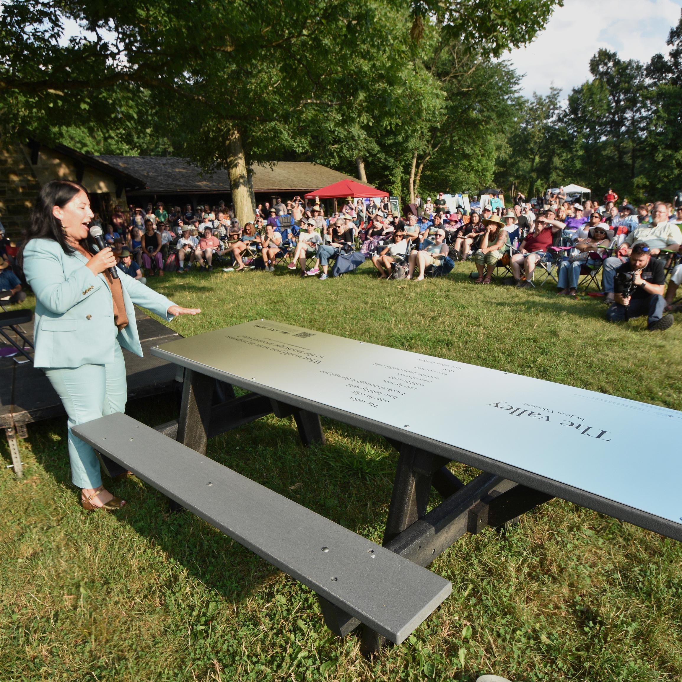 A person speaks to a large crowed in a grassy area next to a picnic table with poem on top.
