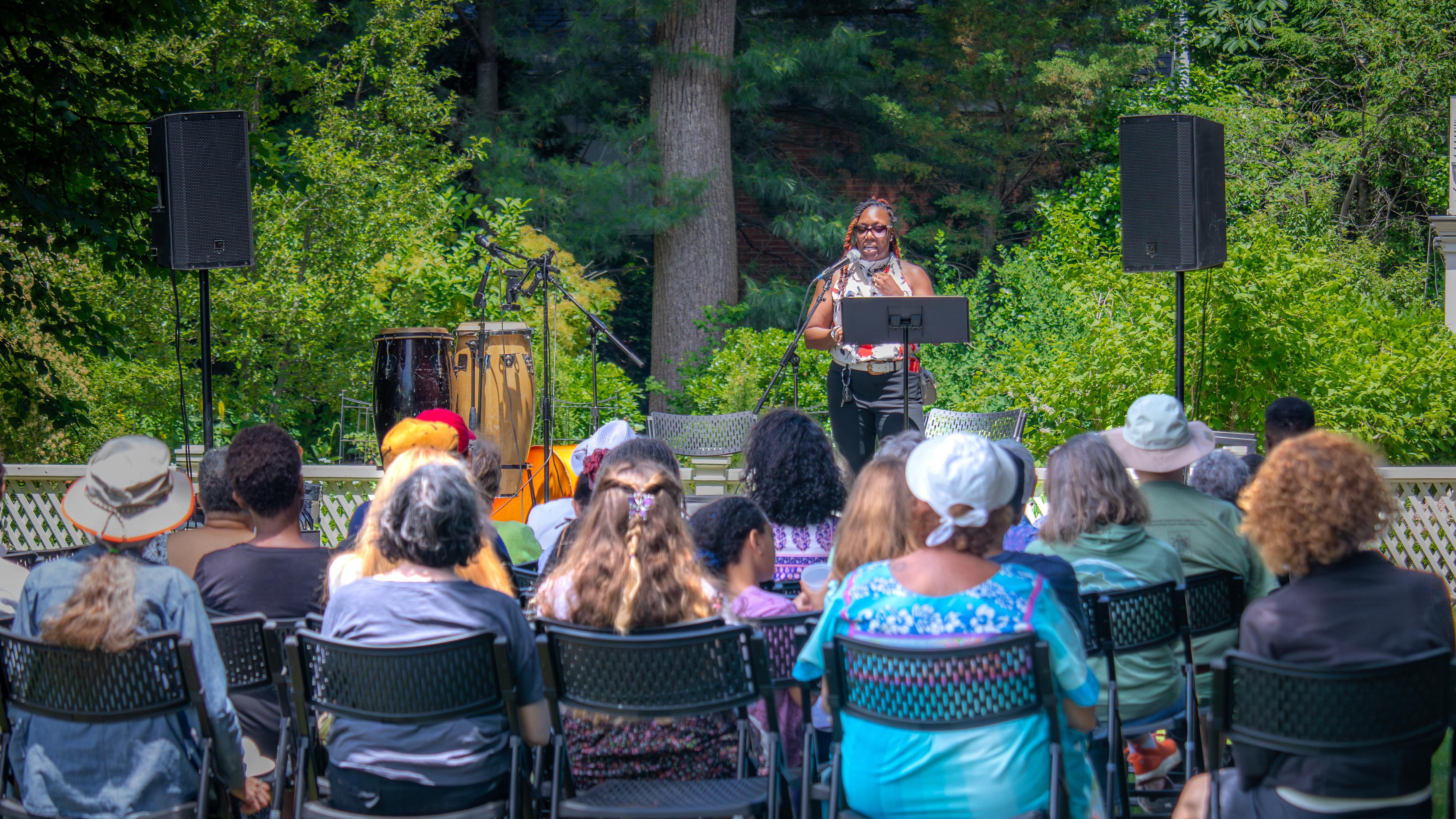 A woman speaks to seated crowd in front of garden.