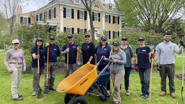 Ten people stand in a garden area in front of a two-story house, holding shovels and tools