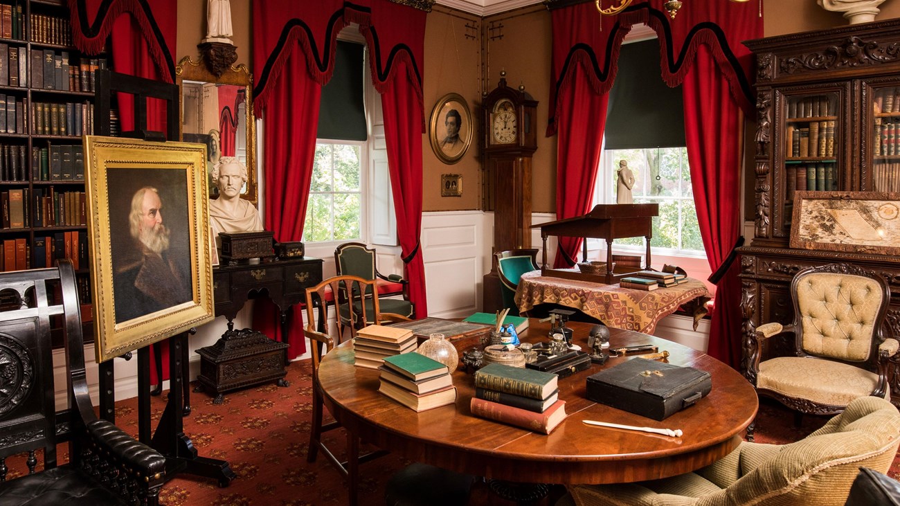 View of a study with a round center table cluttered with books and writing implements.
