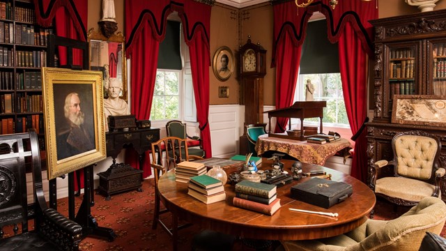 View of a study with a round center table cluttered with books and writing implements.
