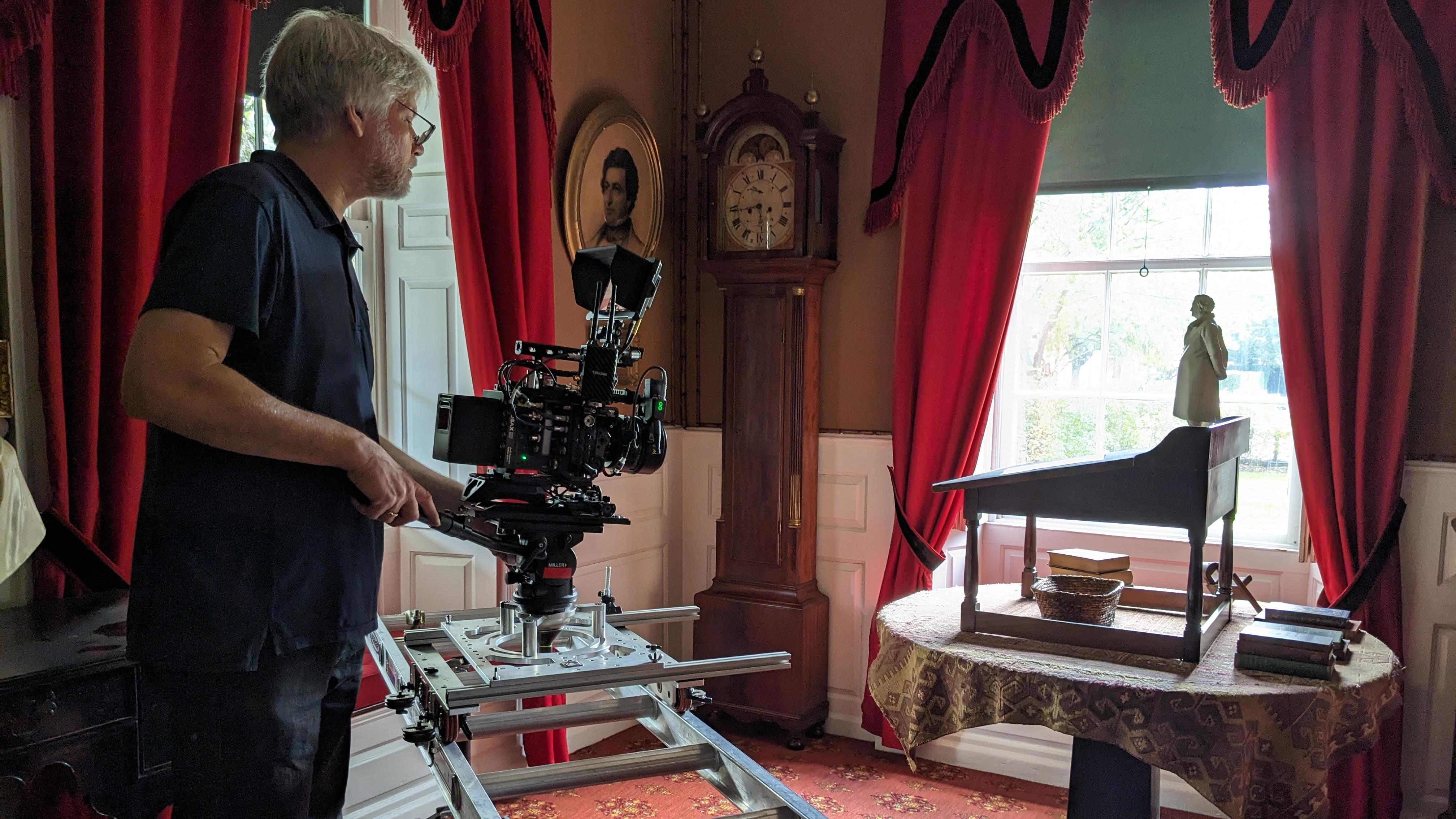 Man with professional videography equipment in a historic study with red curtains and rug.