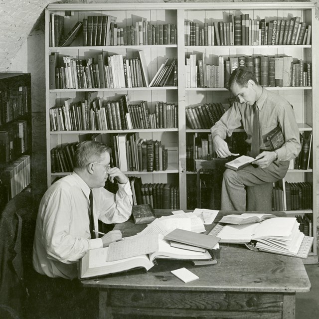 Black and white image of two men looking at documents on table