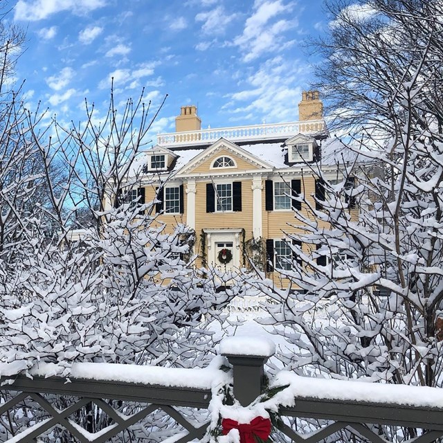 Yellow 3-story mansion with symmetrical facade. Snowy lawn and fence with bows in foreground