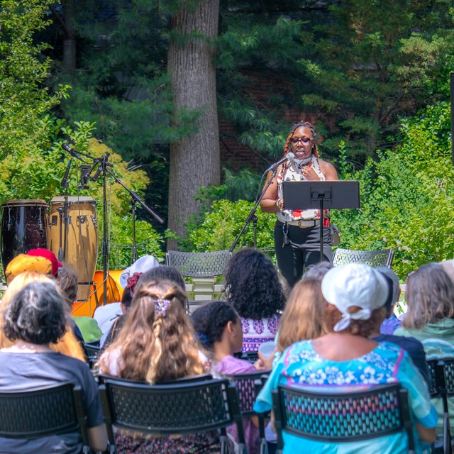 A woman speaks to seated crowd in front of garden.