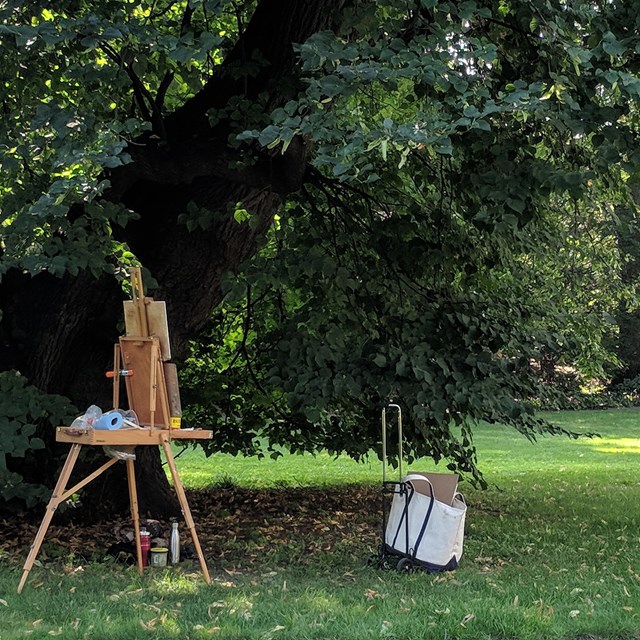 artist's easel set up under large tree, with group of people around second easel on lawn behind