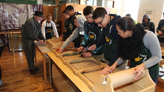 Students wearing aprons work on a mock assembly line while a museum educator guides them