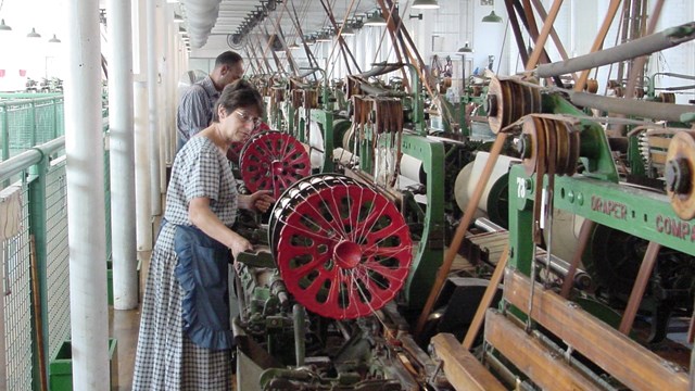 Two people in mill workers' clothing operate power looms