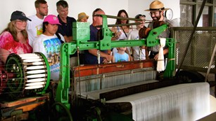 A park ranger explains an old metal weaving machine to a group of visitors of various ages.