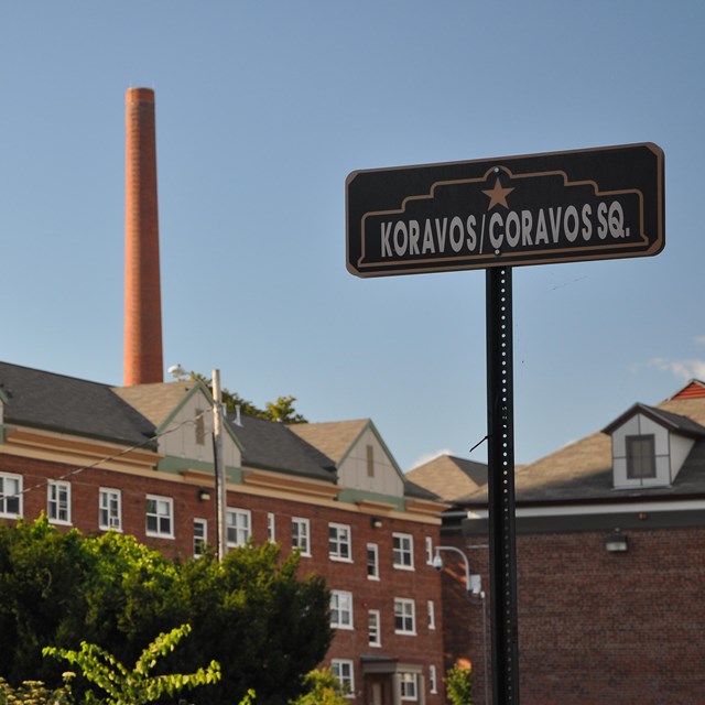 A shot of multiple red brick buildings with a sign in the foreground reading Koravos/Coravos Sq.