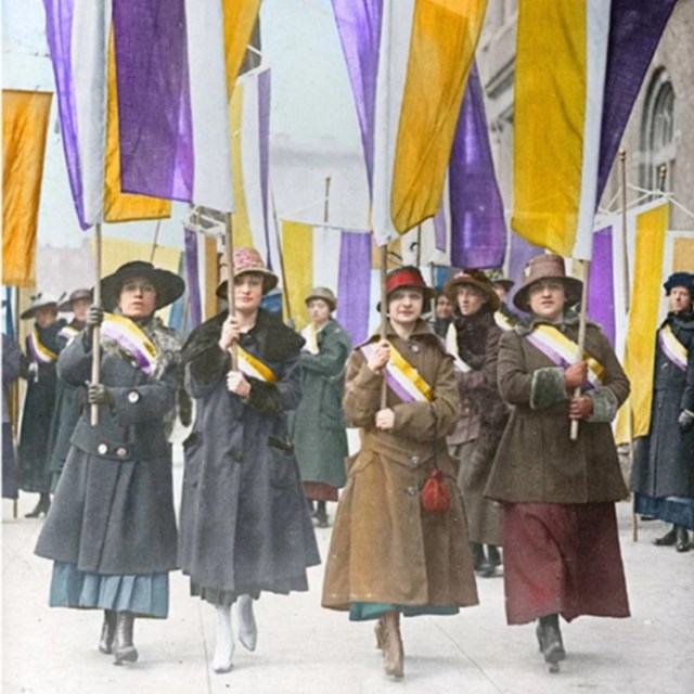 Suffragists bearing their yellow and purple flags and banners marching.