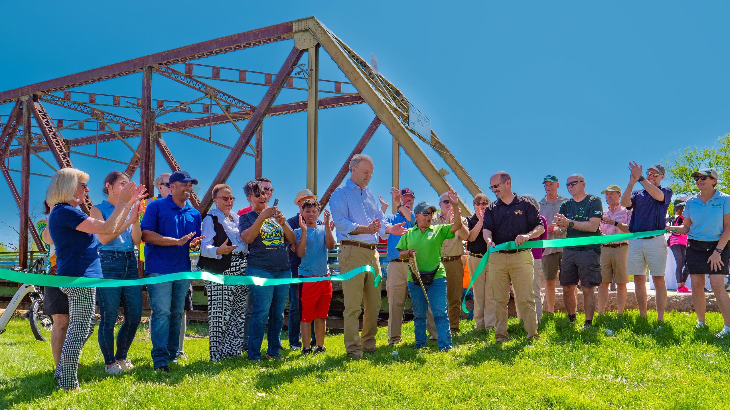 Grassy field with a metal structure and a group of people behind a green ribbon being cut